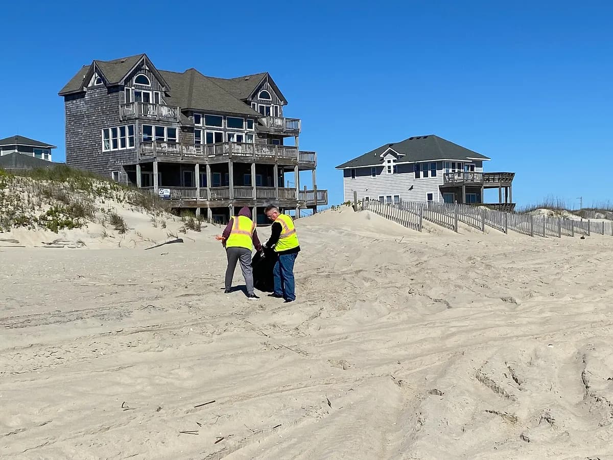 Two volunteers in yellow vests hold a rubbish bag on the sand in front of beach houses.