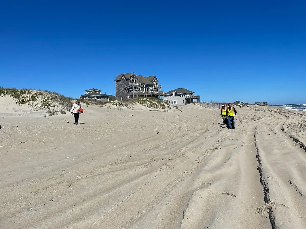 Three volunteers in safety vests walk a wide sandy beach collecting debris under a clear blue sky.